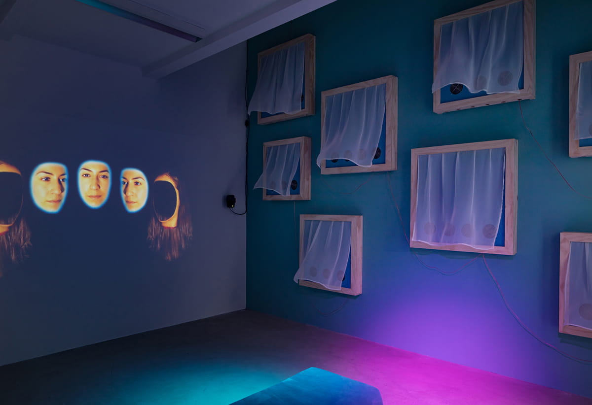 A large film projection to the left of the installation shows two beheaded women whose faces have come off like masks. Blood drips from their necks and the faces turn in the air between them, surrounded by a glowing blue light. The right side of the room is hung with wooden window shaped sculptures, whose curtains are blown out by fans