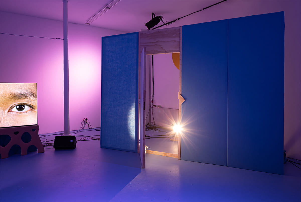 An installation wall made of blue felt stands to the right of the room. In the middle of it a door stands open with a bright light shining through. Behind the door is a wooden sun. To the left of the wall there is a flat screen television showing a close up of a woman’s eyes. The screen is mounted on a carved wooden base, and has a speaker and a pile of cables next to it.